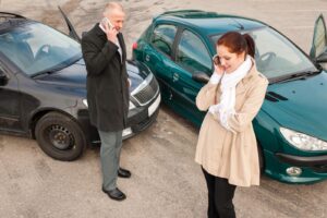 A woman stands in front of a dark green car and a man stands in front of a black car, both on their phones to find out how to get an accident report.